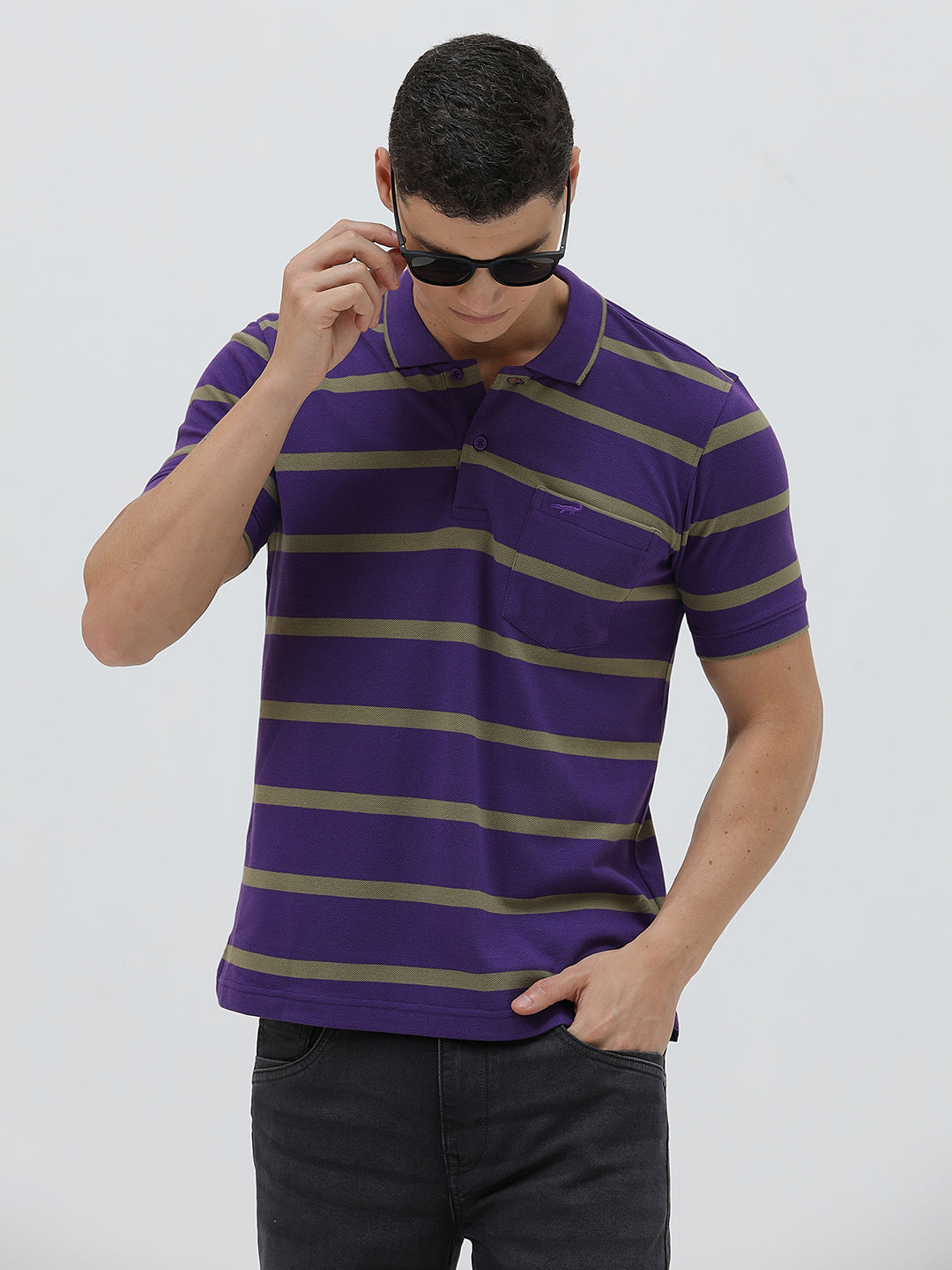 A man wearing a purple short-sleeve polo shirt with horizontal olive green stripes, paired with dark gray pants, standing against a light gray studio background.