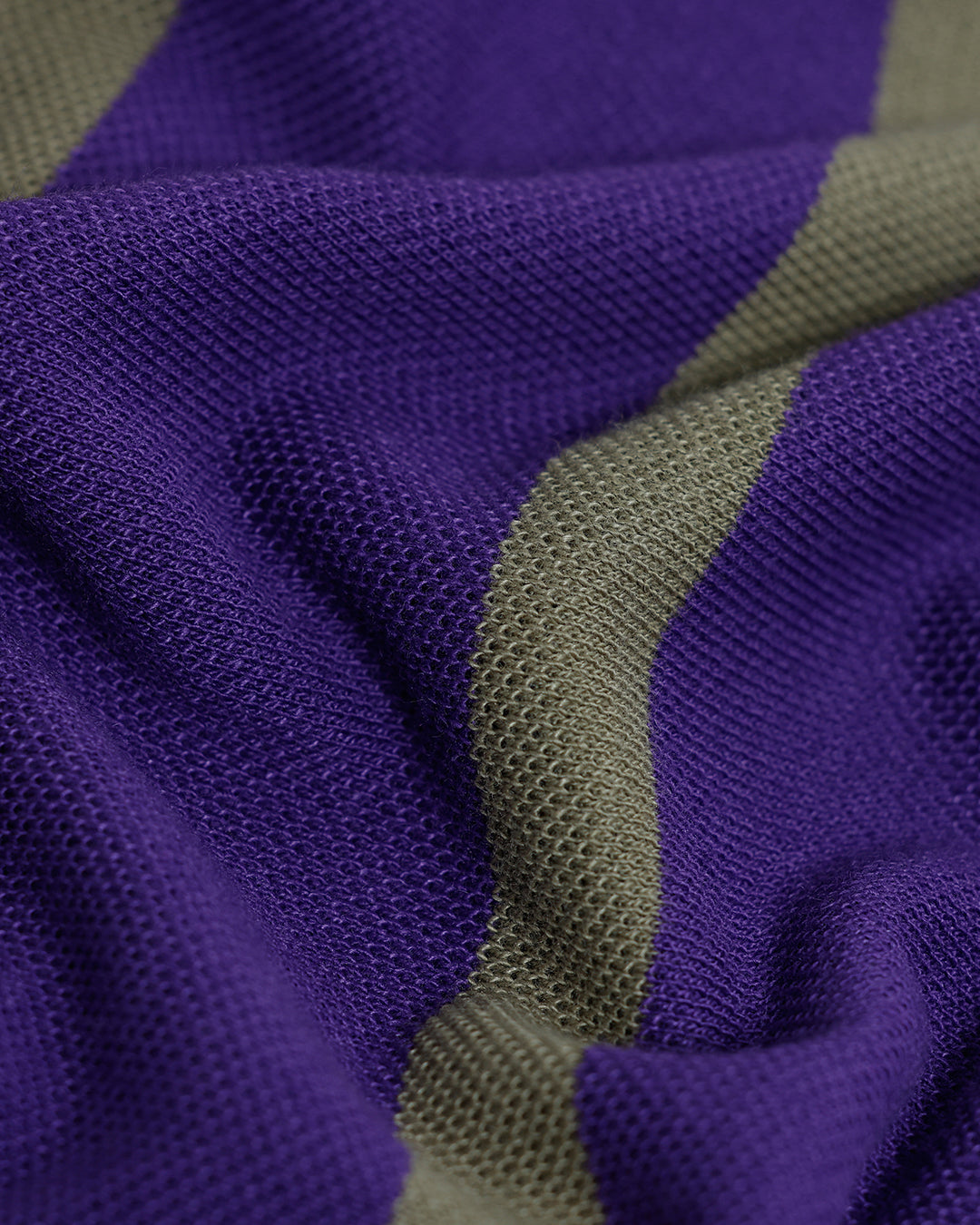 A man wearing a purple short-sleeve polo shirt with horizontal olive green stripes, paired with dark gray pants, standing against a light gray studio background.
