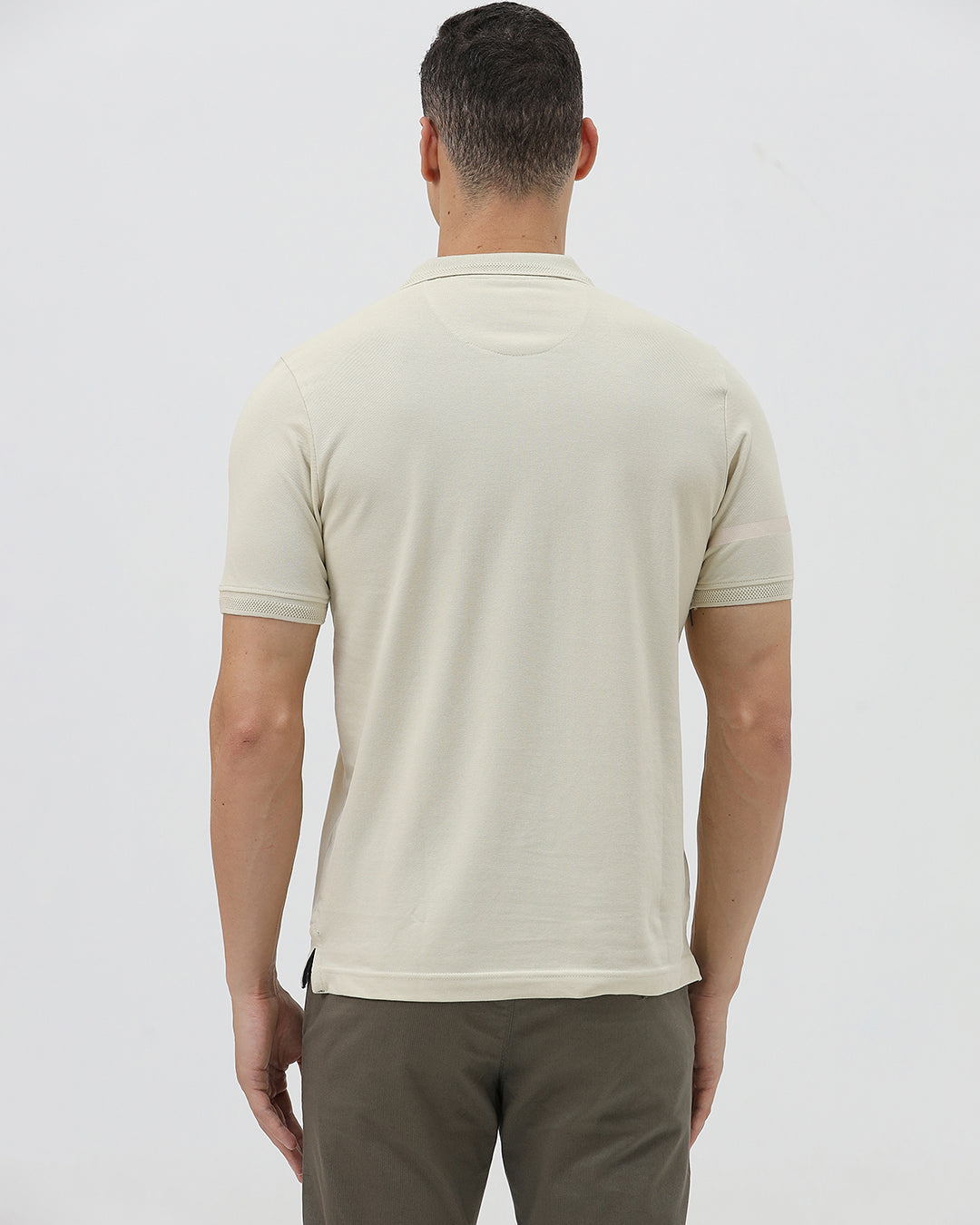 A man wearing a light beige short-sleeve polo shirt with subtle horizontal stripe details and a small crocodile logo on the left chest, paired with olive green trousers, against a plain light gray background.