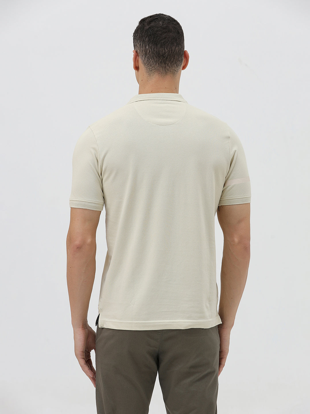 A man wearing a light beige short-sleeve polo shirt with subtle horizontal stripe details and a small crocodile logo on the left chest, paired with olive green trousers, against a plain light gray background.