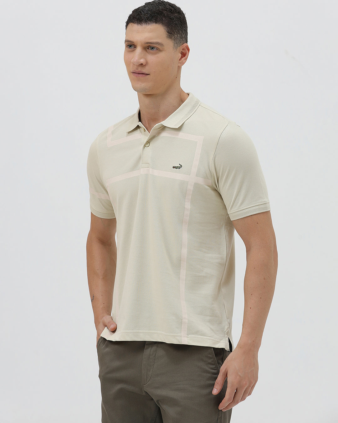 A man wearing a light beige short-sleeve polo shirt with subtle horizontal stripe details and a small crocodile logo on the left chest, paired with olive green trousers, against a plain light gray background.