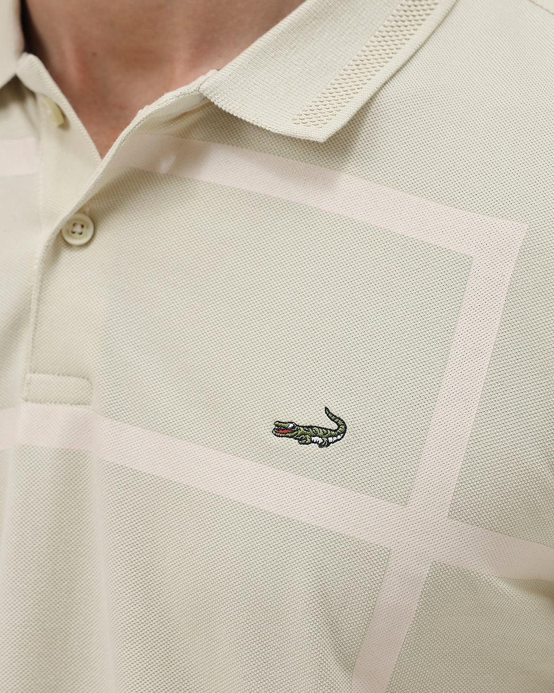 A man wearing a light beige short-sleeve polo shirt with subtle horizontal stripe details and a small crocodile logo on the left chest, paired with olive green trousers, against a plain light gray background.