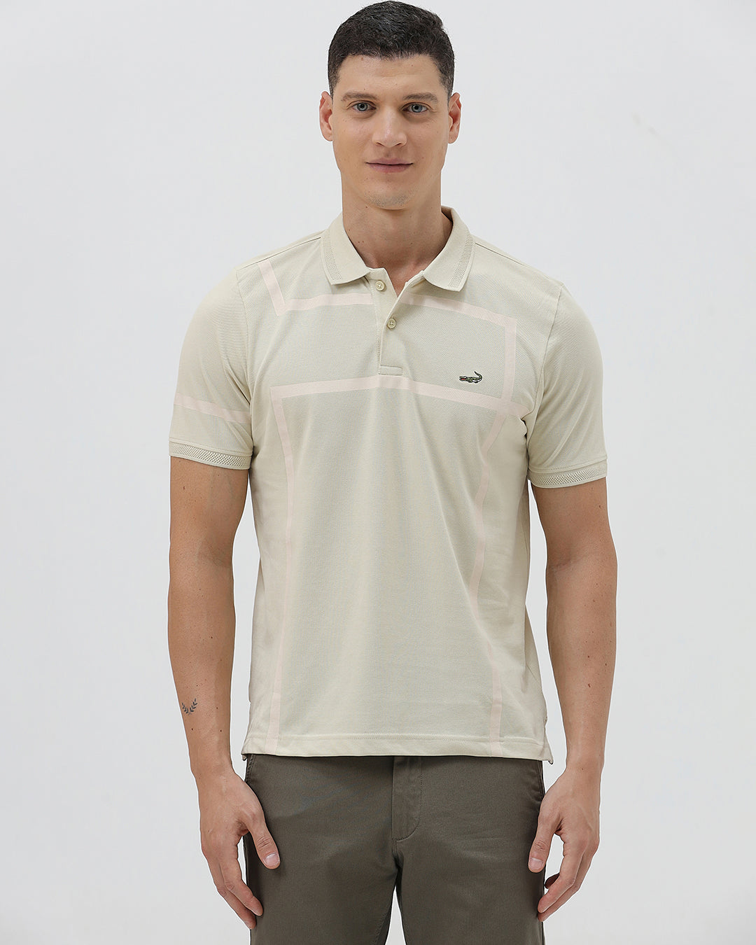 A man wearing a light beige short-sleeve polo shirt with subtle horizontal stripe details and a small crocodile logo on the left chest, paired with olive green trousers, against a plain light gray background.