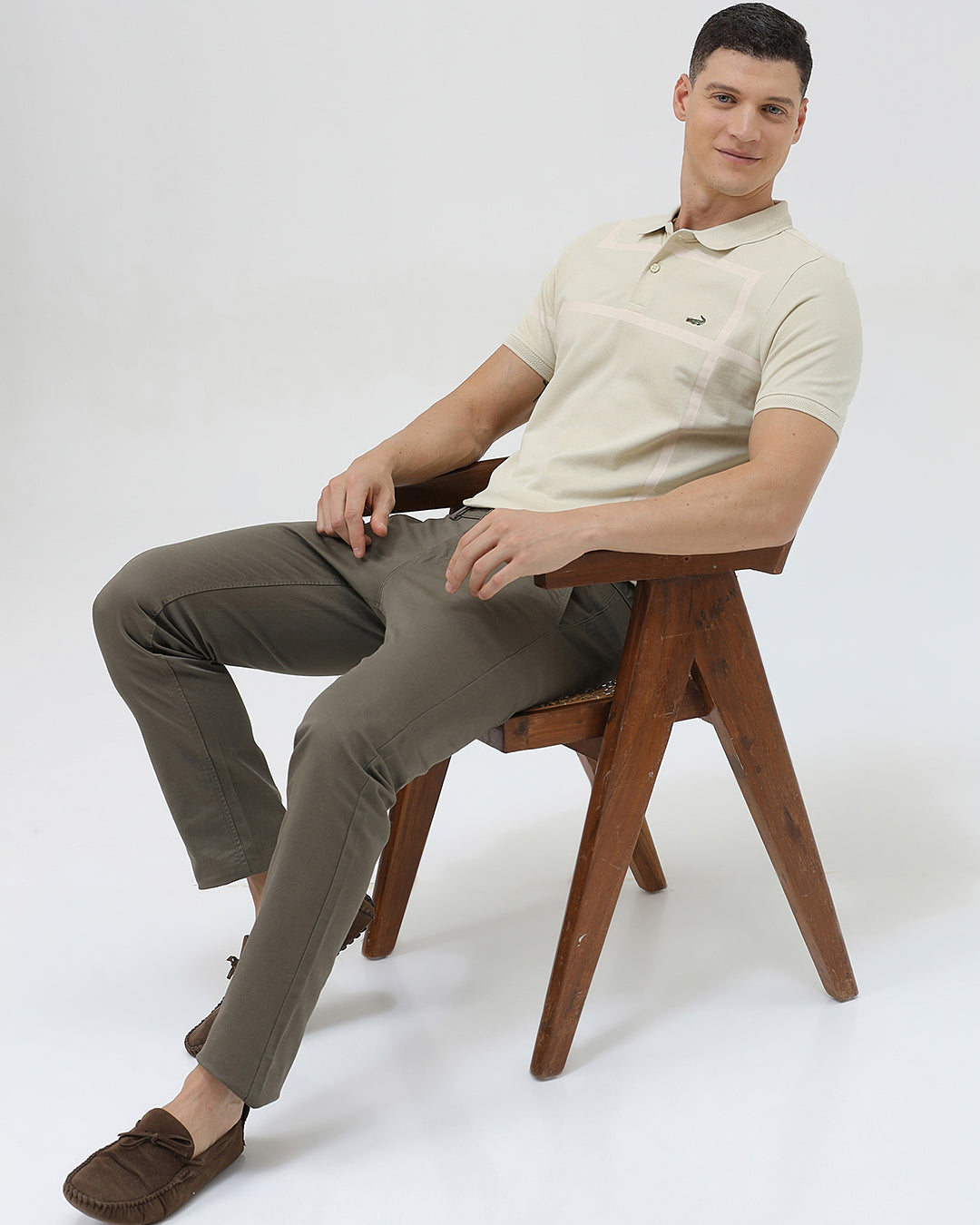 A man wearing a light beige short-sleeve polo shirt with subtle horizontal stripe details and a small crocodile logo on the left chest, paired with olive green trousers, against a plain light gray background.
