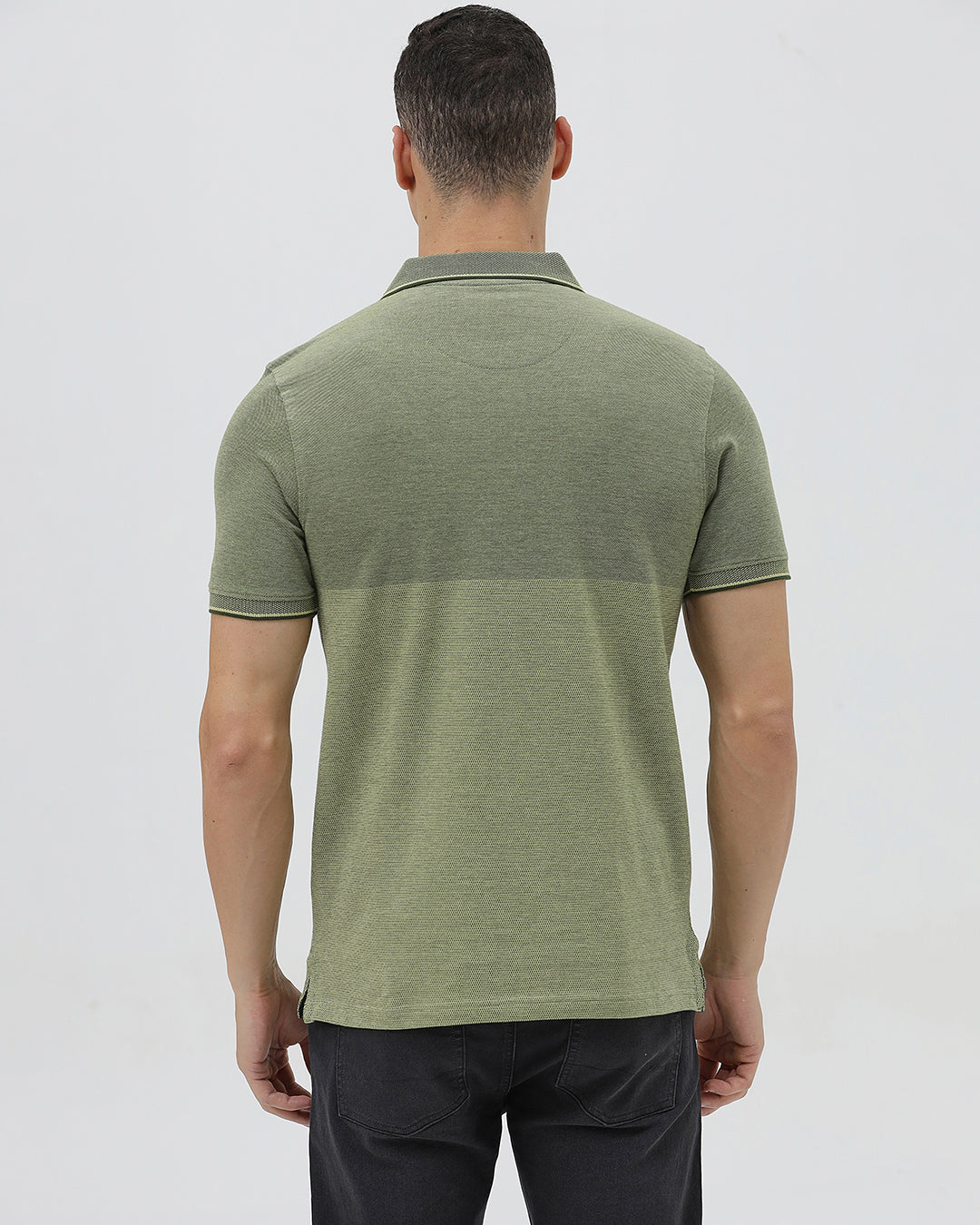 A man wearing a short-sleeve polo shirt in olive green with a subtle horizontal color-blocked stripe and a small gold crest logo on the left chest, paired with dark pants, against a light gray studio background.