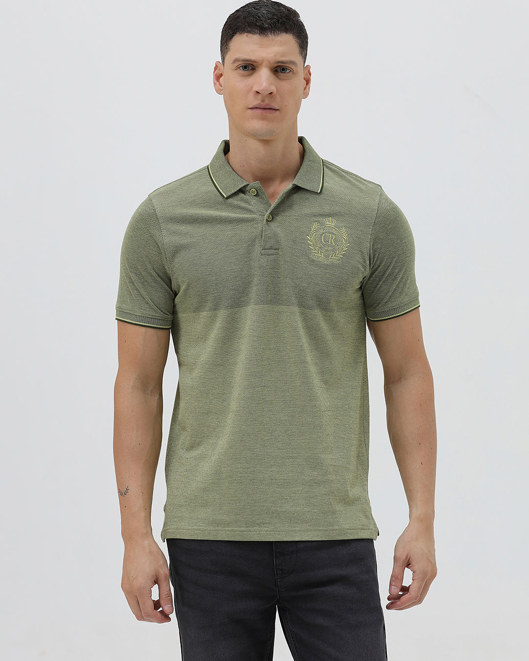 A man wearing a short-sleeve polo shirt in olive green with a subtle horizontal color-blocked stripe and a small gold crest logo on the left chest, paired with dark pants, against a light gray studio background.