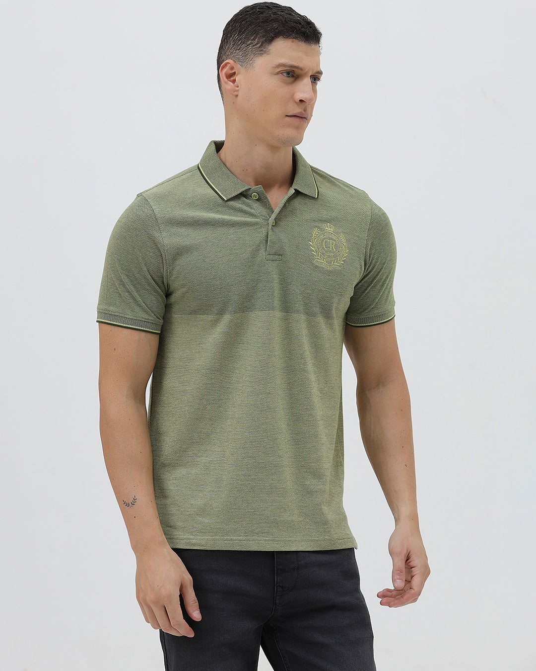 A man wearing a short-sleeve polo shirt in olive green with a subtle horizontal color-blocked stripe and a small gold crest logo on the left chest, paired with dark pants, against a light gray studio background.
