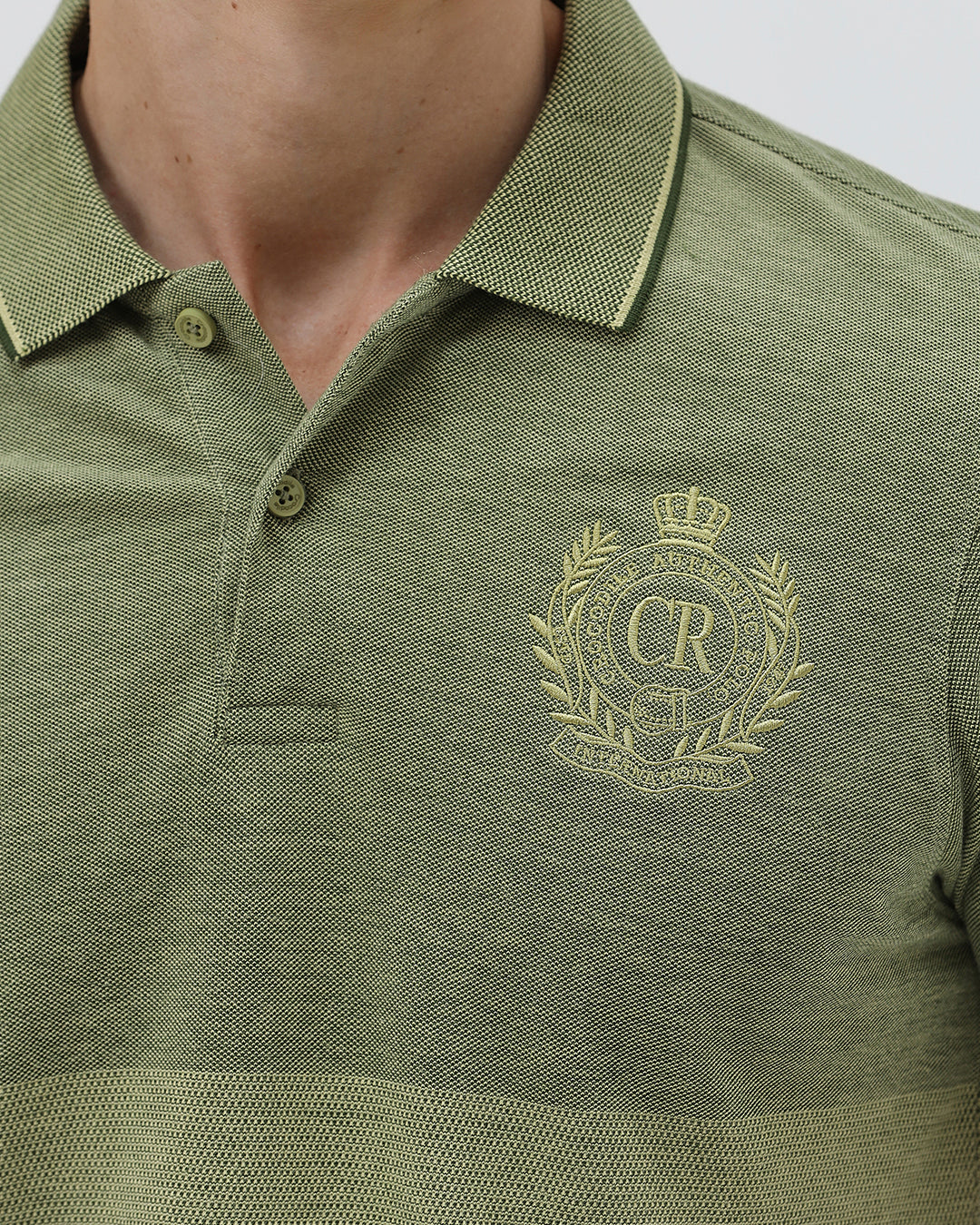 A man wearing a short-sleeve polo shirt in olive green with a subtle horizontal color-blocked stripe and a small gold crest logo on the left chest, paired with dark pants, against a light gray studio background.