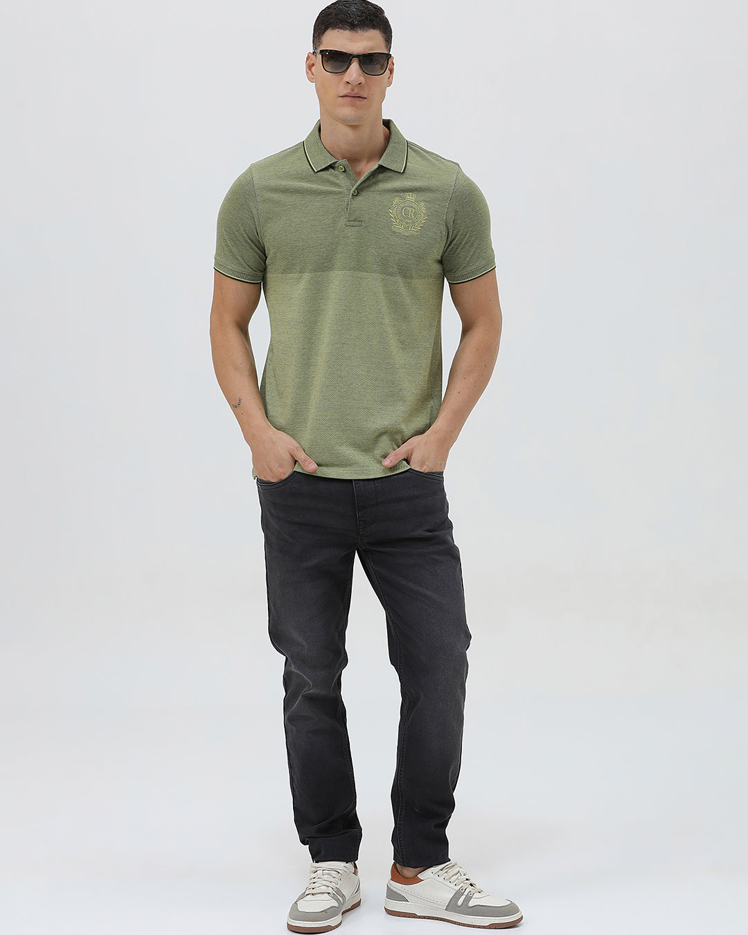 A man wearing a short-sleeve polo shirt in olive green with a subtle horizontal color-blocked stripe and a small gold crest logo on the left chest, paired with dark pants, against a light gray studio background.