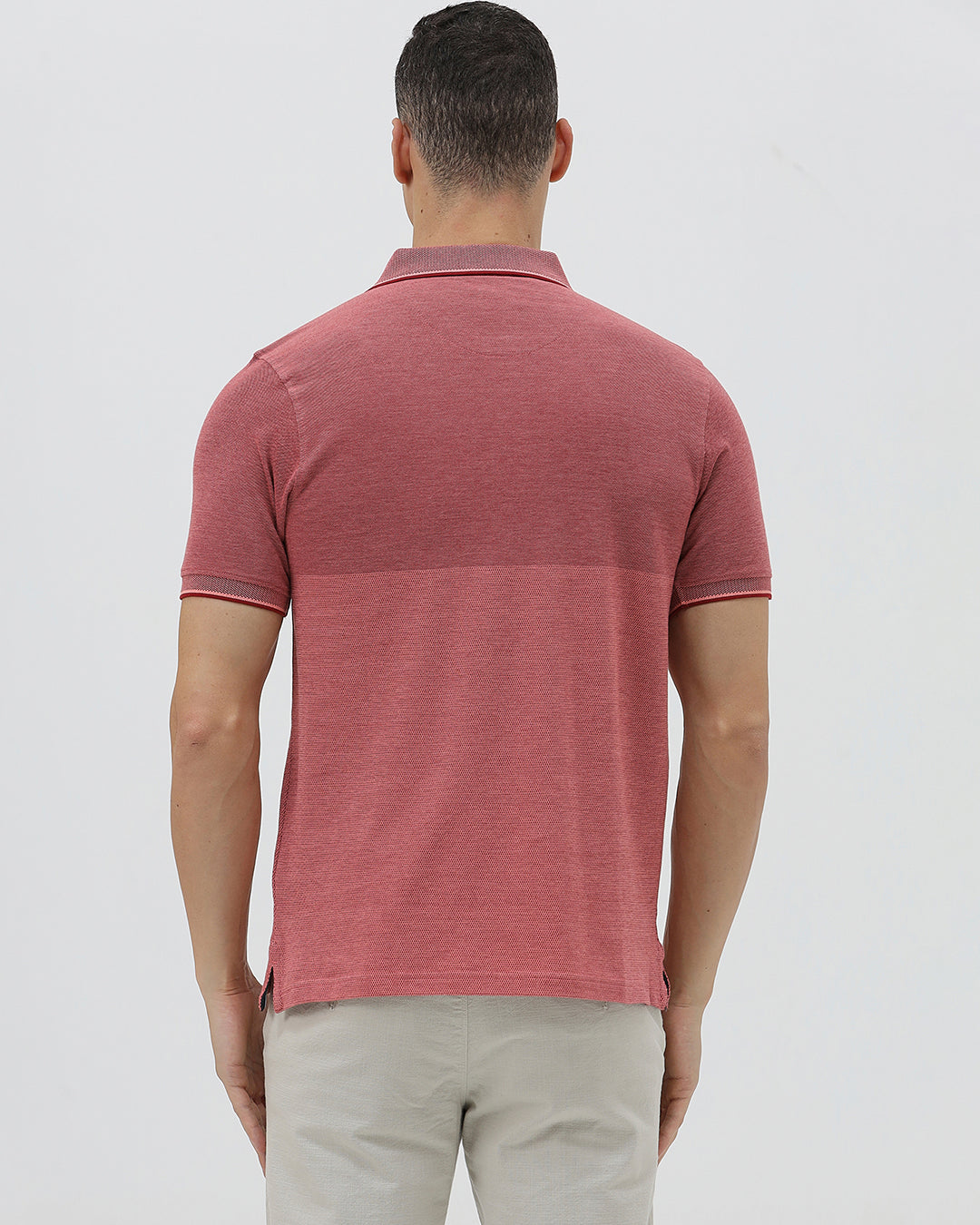 A man wearing a heathered dusty rose pink polo shirt with a tonal embroidered crest on the left chest, paired with light beige trousers, against a plain light gray background.