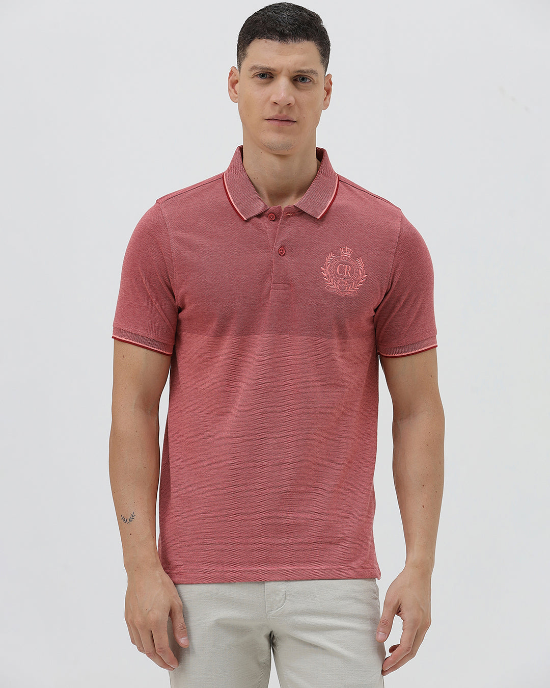 A man wearing a heathered dusty rose pink polo shirt with a tonal embroidered crest on the left chest, paired with light beige trousers, against a plain light gray background.