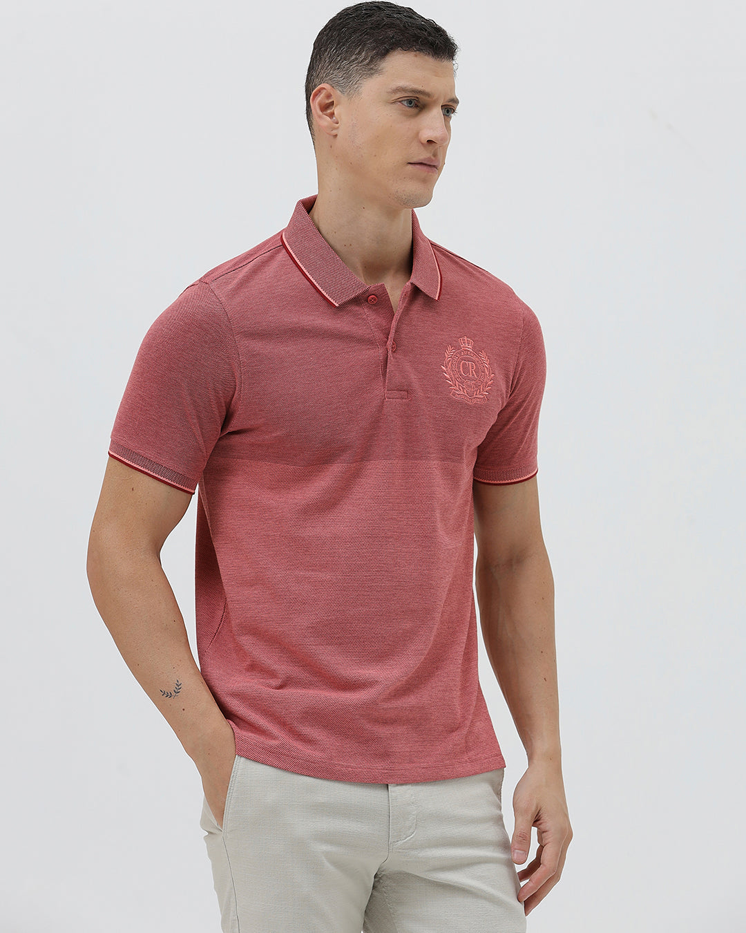 A man wearing a heathered dusty rose pink polo shirt with a tonal embroidered crest on the left chest, paired with light beige trousers, against a plain light gray background.