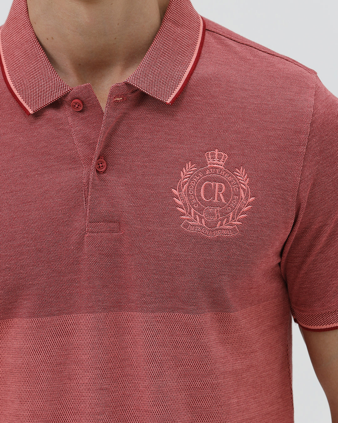 A man wearing a heathered dusty rose pink polo shirt with a tonal embroidered crest on the left chest, paired with light beige trousers, against a plain light gray background.