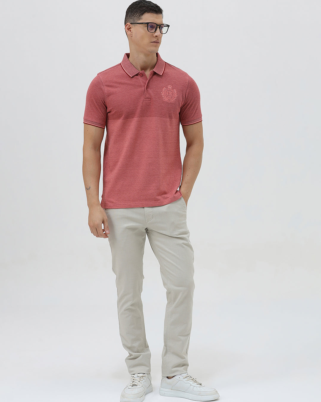 A man wearing a heathered dusty rose pink polo shirt with a tonal embroidered crest on the left chest, paired with light beige trousers, against a plain light gray background.