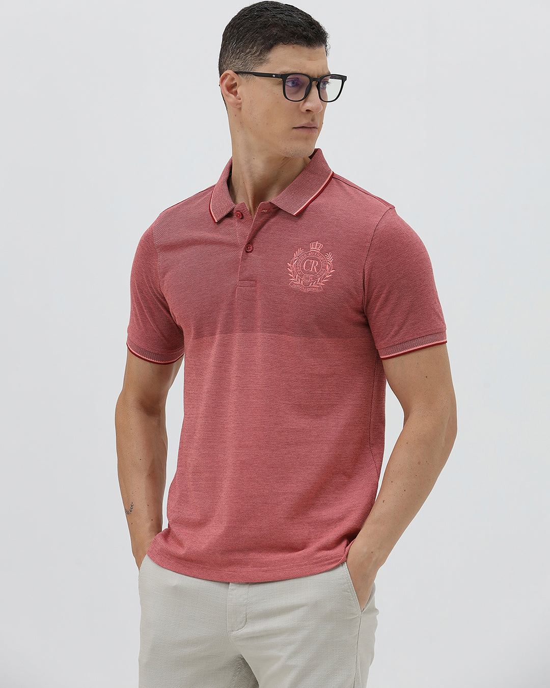 A man wearing a heathered dusty rose pink polo shirt with a tonal embroidered crest on the left chest, paired with light beige trousers, against a plain light gray background.