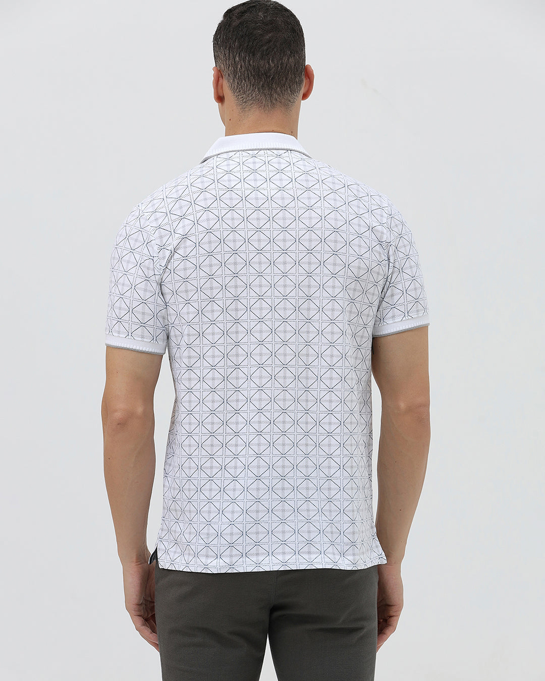 A man wearing a white short-sleeve polo shirt with a gray geometric pattern, paired with dark gray trousers, standing against a light gray studio background.