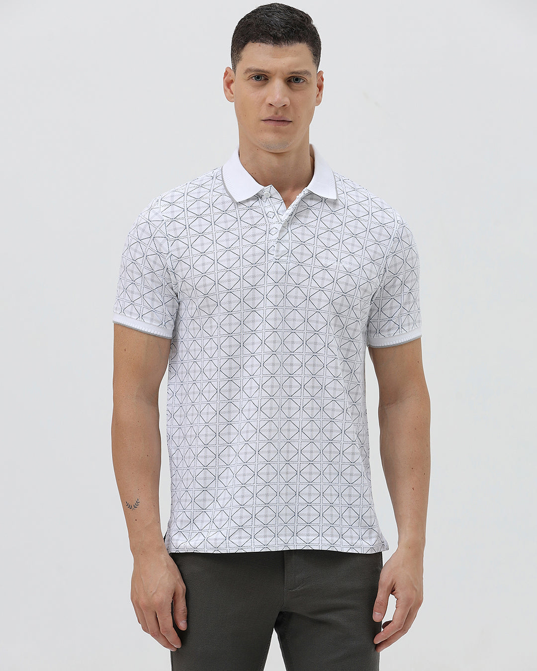 A man wearing a white short-sleeve polo shirt with a gray geometric pattern, paired with dark gray trousers, standing against a light gray studio background.