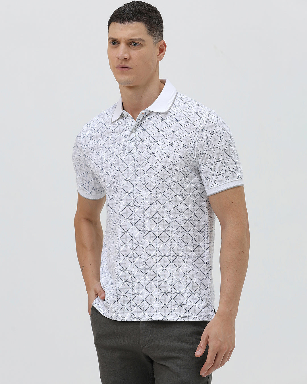 A man wearing a white short-sleeve polo shirt with a gray geometric pattern, paired with dark gray trousers, standing against a light gray studio background.