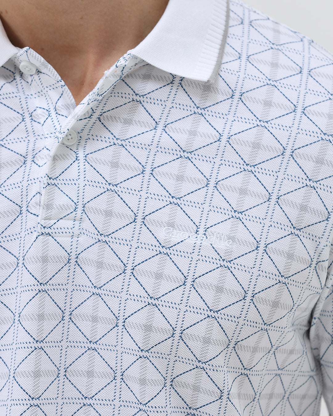 A man wearing a white short-sleeve polo shirt with a gray geometric pattern, paired with dark gray trousers, standing against a light gray studio background.
