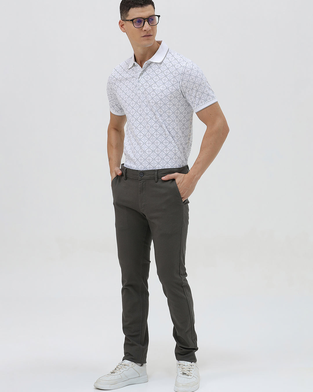 A man wearing a white short-sleeve polo shirt with a gray geometric pattern, paired with dark gray trousers, standing against a light gray studio background.