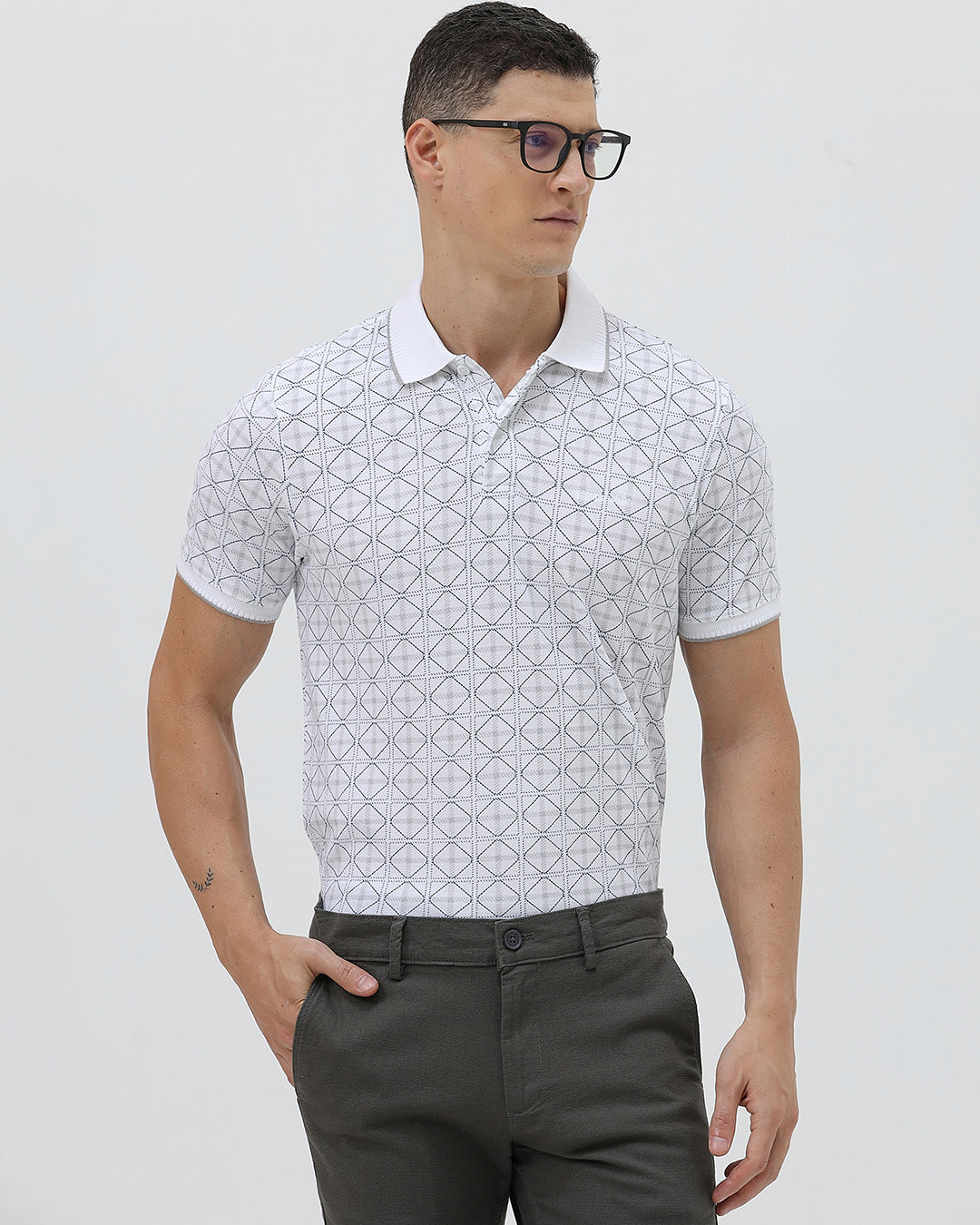 A man wearing a white short-sleeve polo shirt with a gray geometric pattern, paired with dark gray trousers, standing against a light gray studio background.
