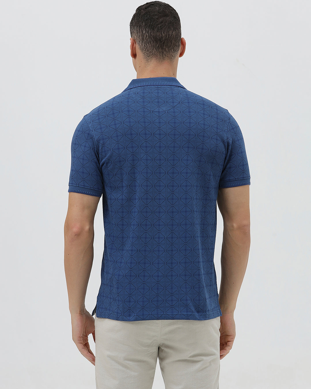 A man wearing a blue short-sleeve polo shirt with a subtle geometric pattern, paired with beige trousers, standing against a light gray studio background.