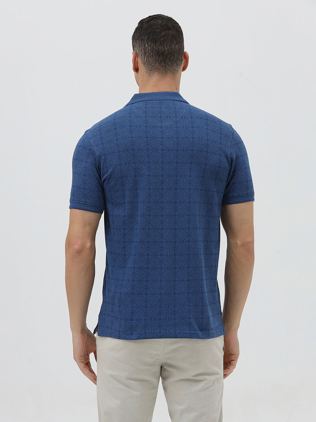 A man wearing a blue short-sleeve polo shirt with a subtle geometric pattern, paired with beige trousers, standing against a light gray studio background.
