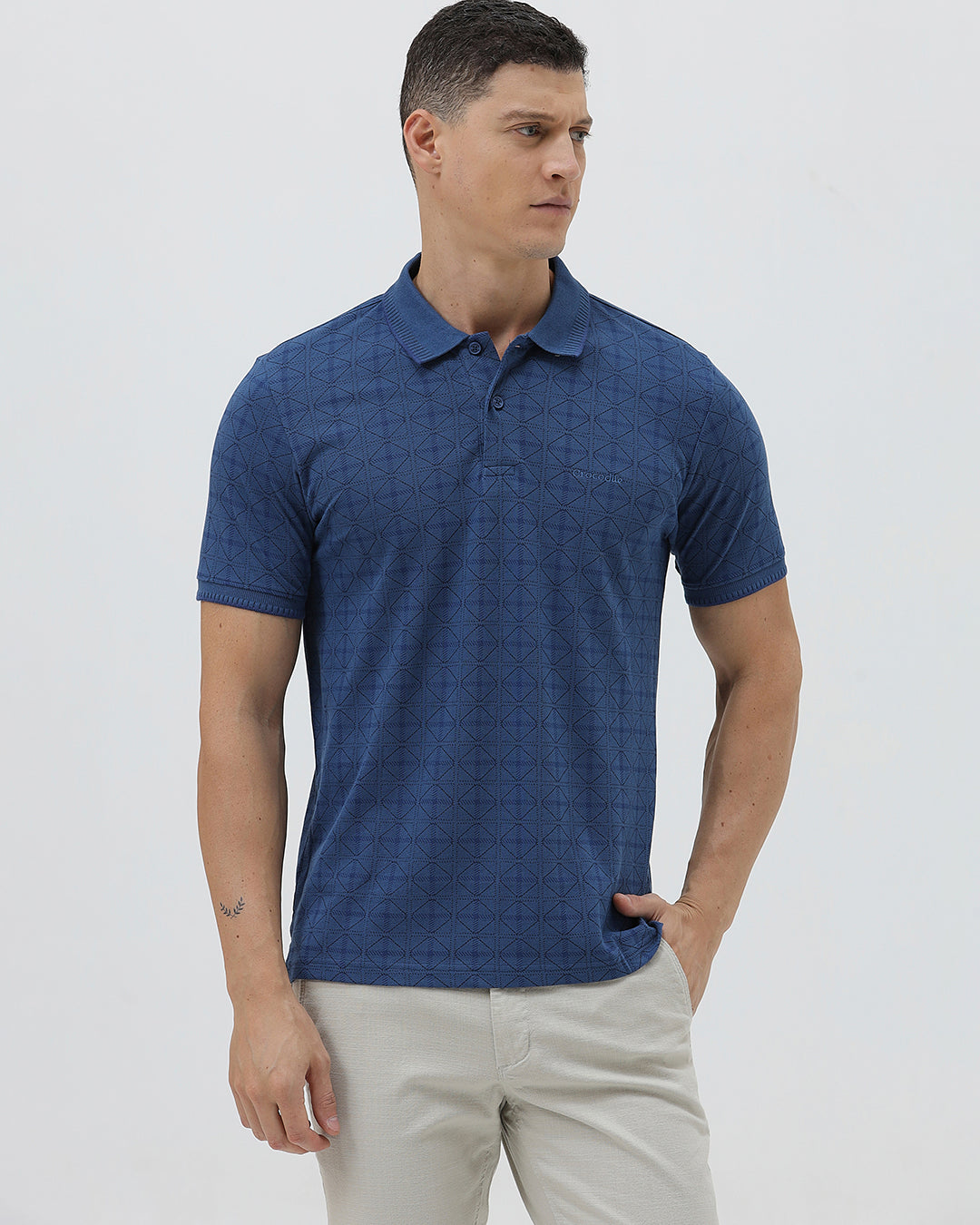 A man wearing a blue short-sleeve polo shirt with a subtle geometric pattern, paired with beige trousers, standing against a light gray studio background.
