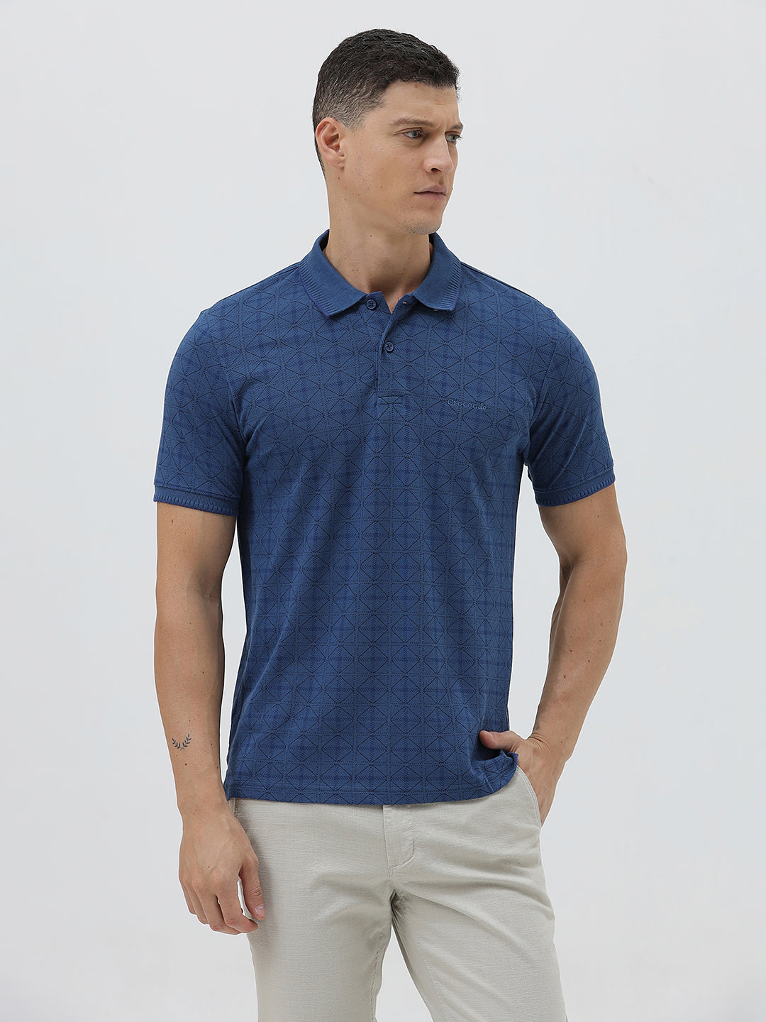 A man wearing a blue short-sleeve polo shirt with a subtle geometric pattern, paired with beige trousers, standing against a light gray studio background.
