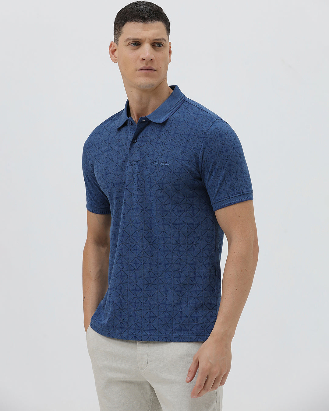 A man wearing a blue short-sleeve polo shirt with a subtle geometric pattern, paired with beige trousers, standing against a light gray studio background.