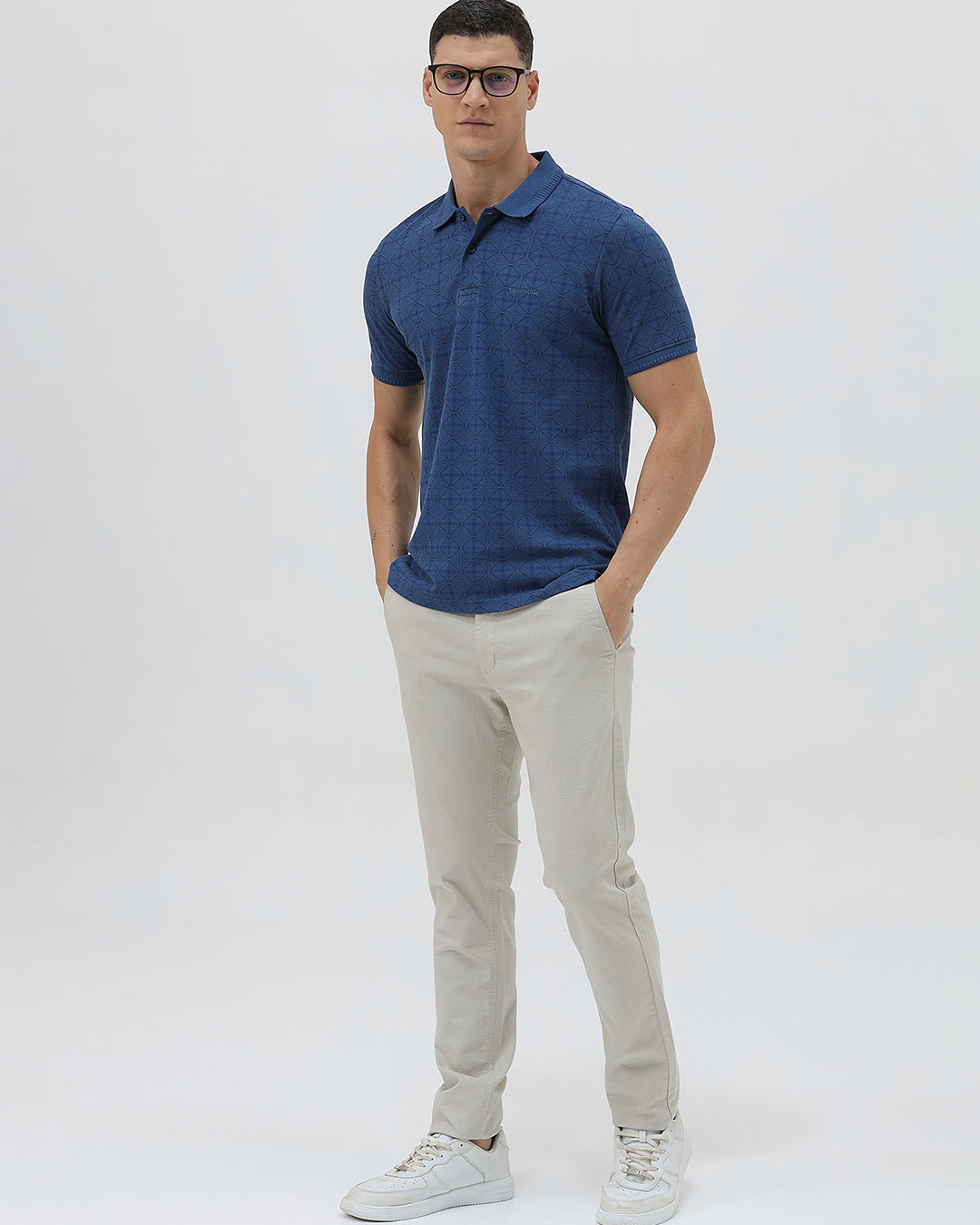 A man wearing a blue short-sleeve polo shirt with a subtle geometric pattern, paired with beige trousers, standing against a light gray studio background.