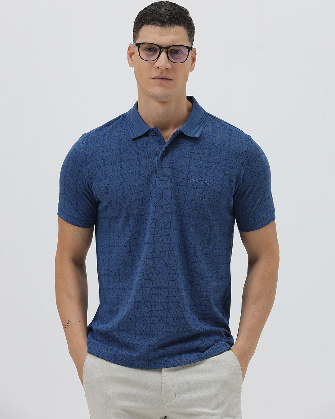 A man wearing a blue short-sleeve polo shirt with a subtle geometric pattern, paired with beige trousers, standing against a light gray studio background.