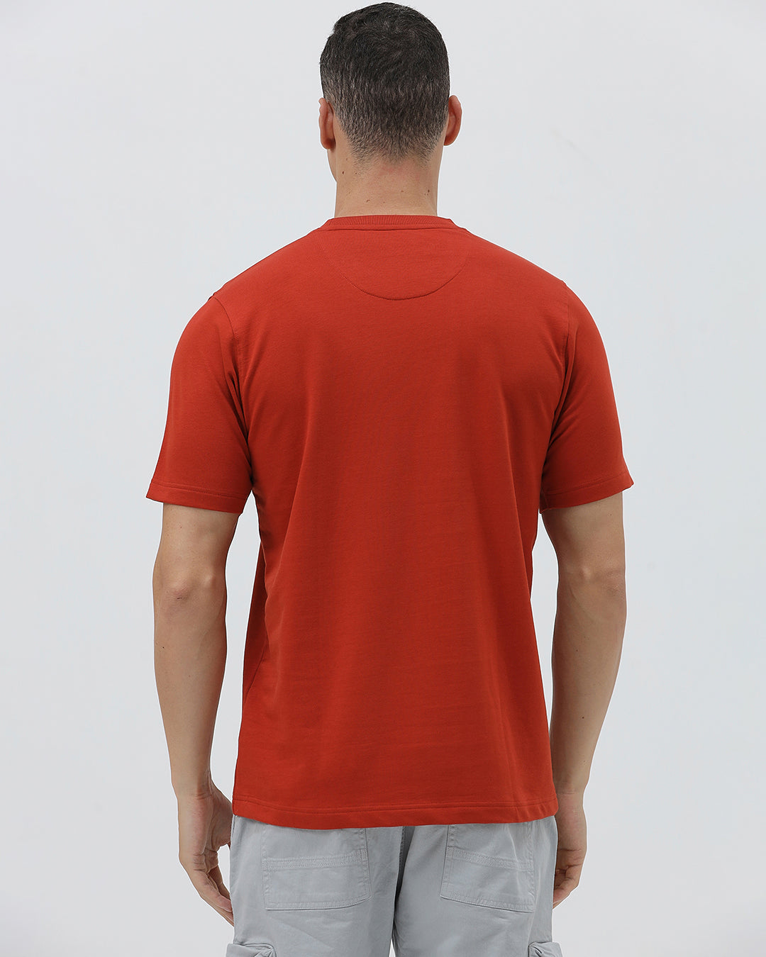 A man wearing a short-sleeve crewneck t-shirt in a muted brick red color, paired with light gray cargo pants, standing against a light gray studio background.