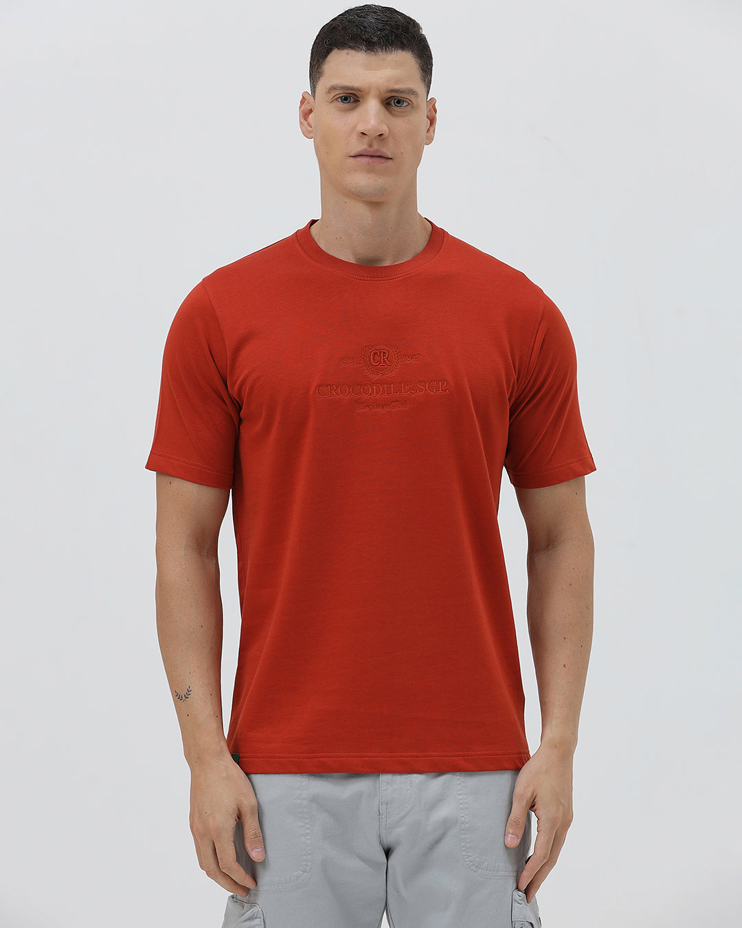 A man wearing a short-sleeve crewneck t-shirt in a muted brick red color, paired with light gray cargo pants, standing against a light gray studio background.