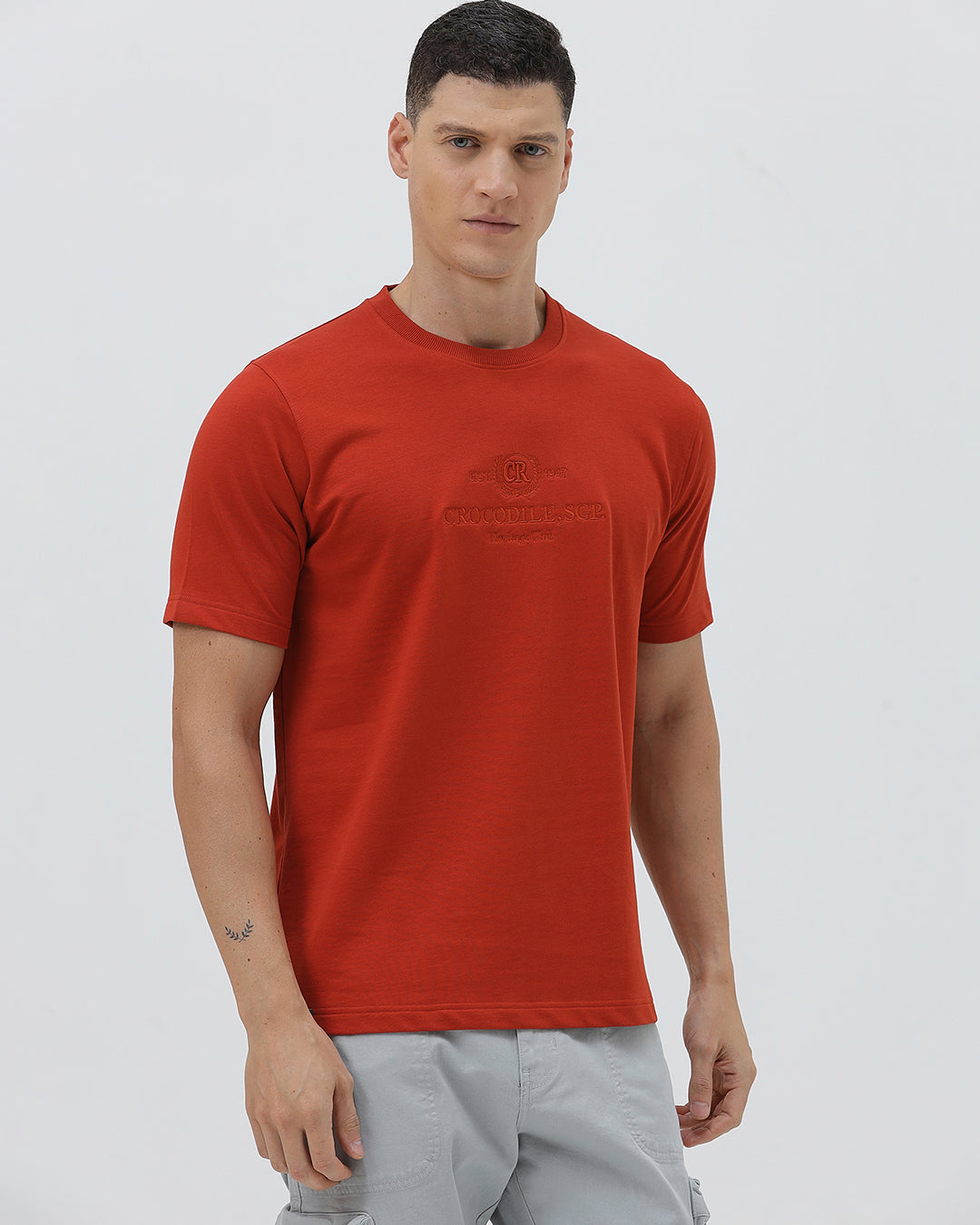 A man wearing a short-sleeve crewneck t-shirt in a muted brick red color, paired with light gray cargo pants, standing against a light gray studio background.