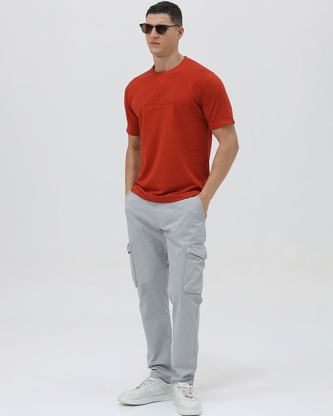 A man wearing a short-sleeve crewneck t-shirt in a muted brick red color, paired with light gray cargo pants, standing against a light gray studio background.