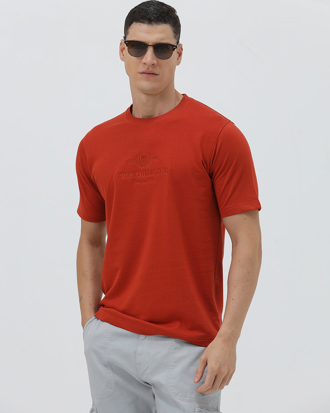A man wearing a short-sleeve crewneck t-shirt in a muted brick red color, paired with light gray cargo pants, standing against a light gray studio background.