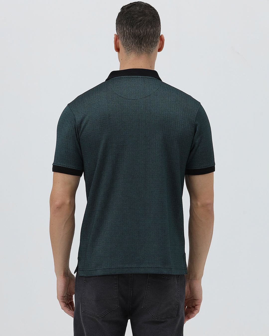 A man wearing a dark green short-sleeve polo shirt with a black collar and black sleeve trim, paired with dark gray jeans, against a light gray studio background.