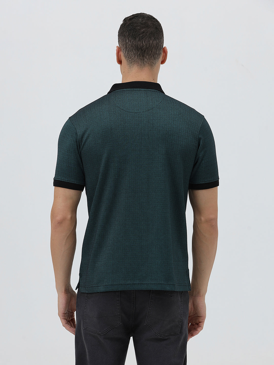 A man wearing a dark green short-sleeve polo shirt with a black collar and black sleeve trim, paired with dark gray jeans, against a light gray studio background.