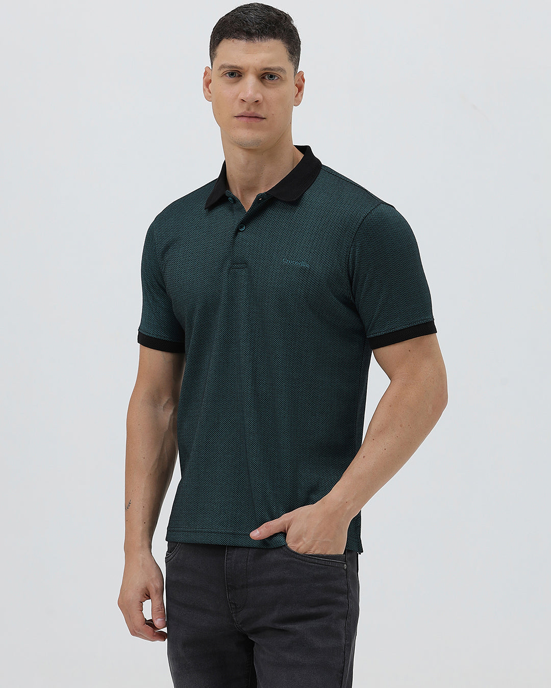 A man wearing a dark green short-sleeve polo shirt with a black collar and black sleeve trim, paired with dark gray jeans, against a light gray studio background.