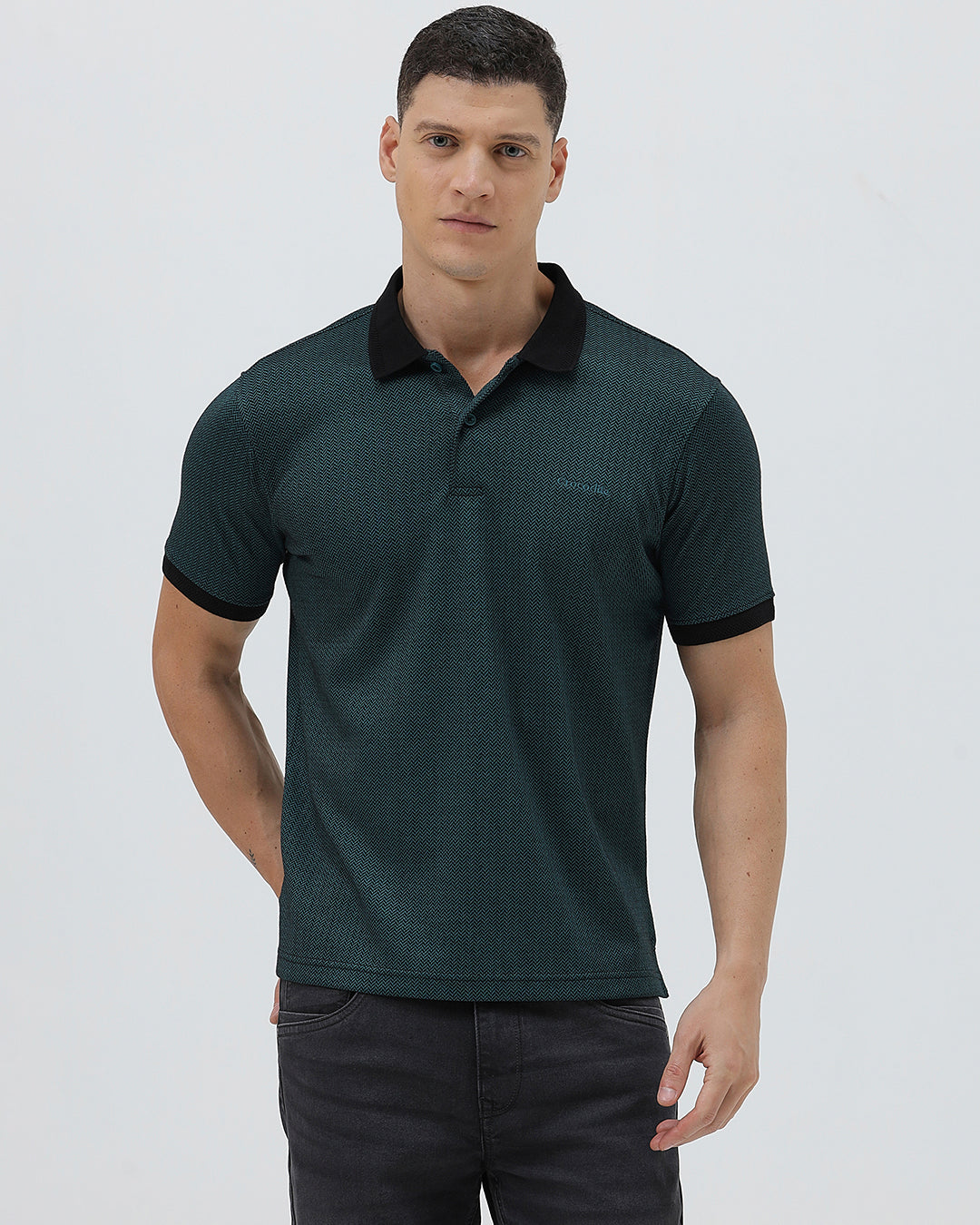 A man wearing a dark green short-sleeve polo shirt with a black collar and black sleeve trim, paired with dark gray jeans, against a light gray studio background.