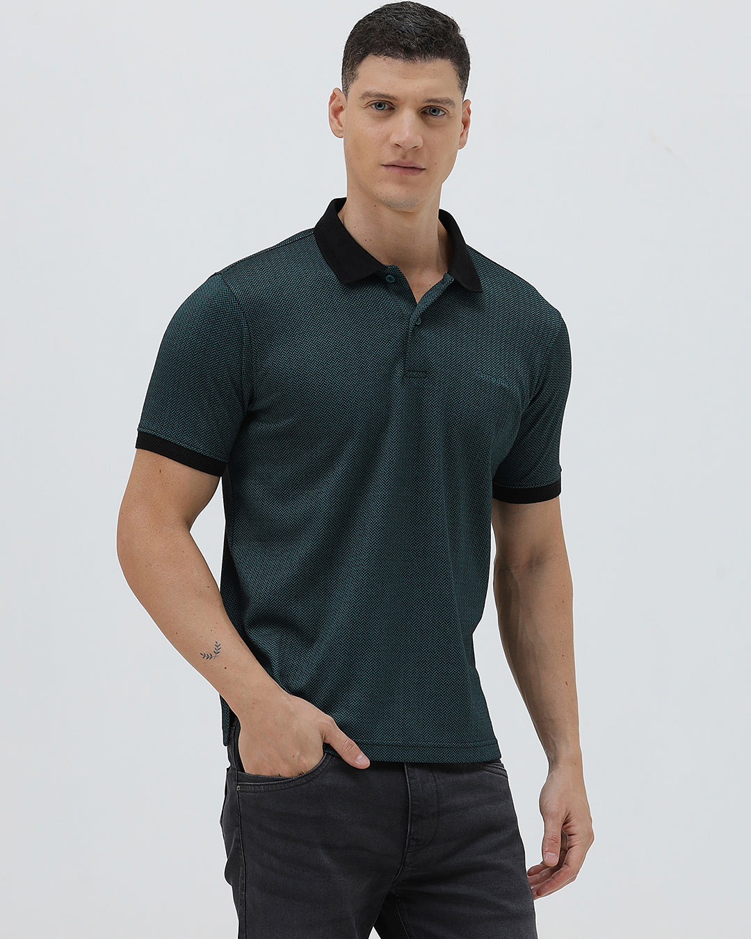 A man wearing a dark green short-sleeve polo shirt with a black collar and black sleeve trim, paired with dark gray jeans, against a light gray studio background.