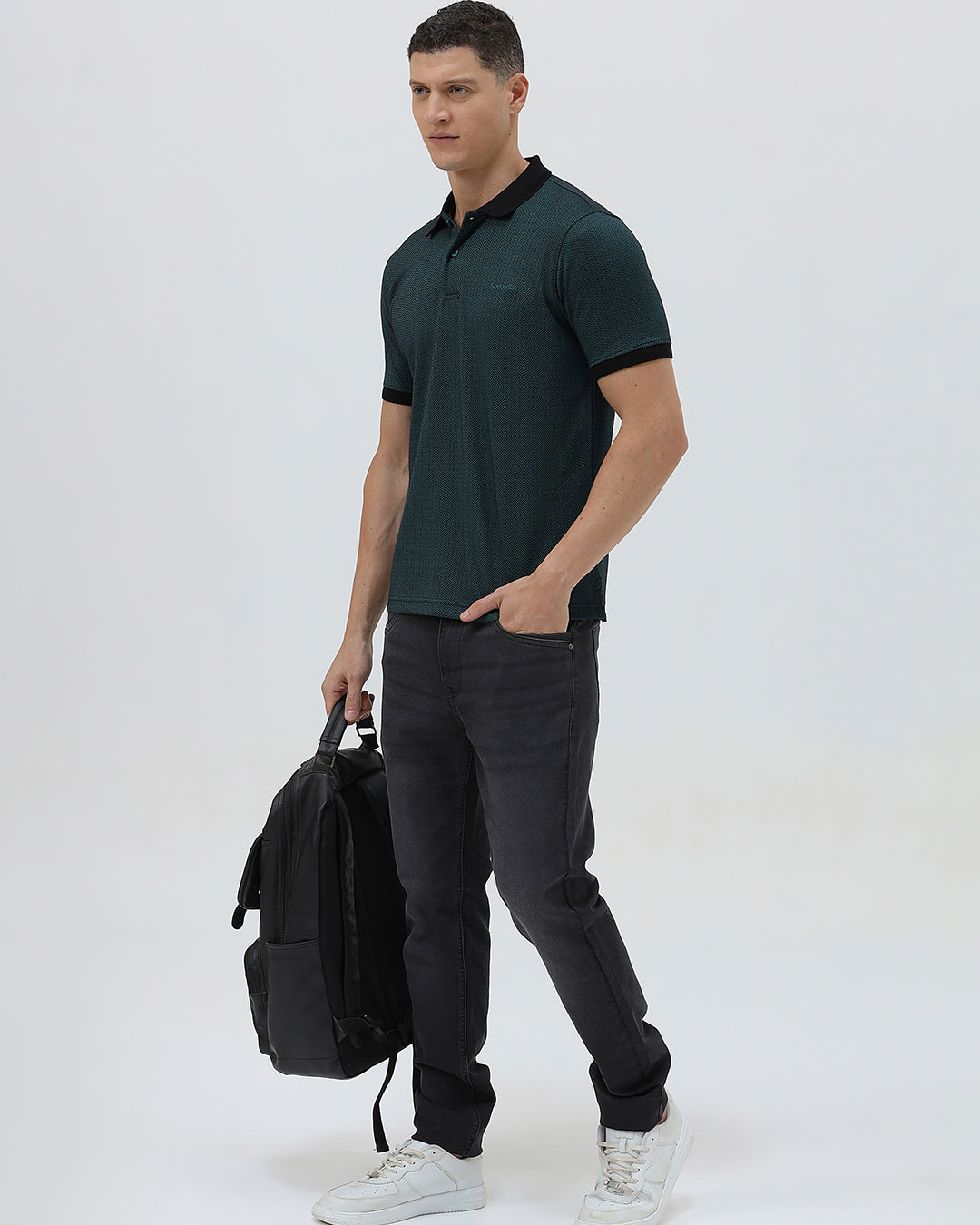 A man wearing a dark green short-sleeve polo shirt with a black collar and black sleeve trim, paired with dark gray jeans, against a light gray studio background.