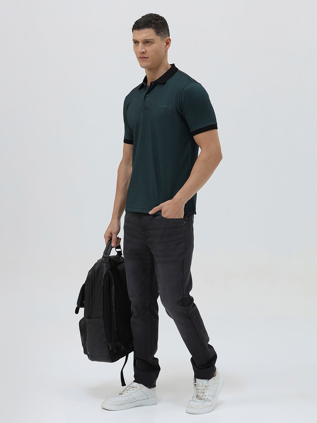 A man wearing a dark green short-sleeve polo shirt with a black collar and black sleeve trim, paired with dark gray jeans, against a light gray studio background.