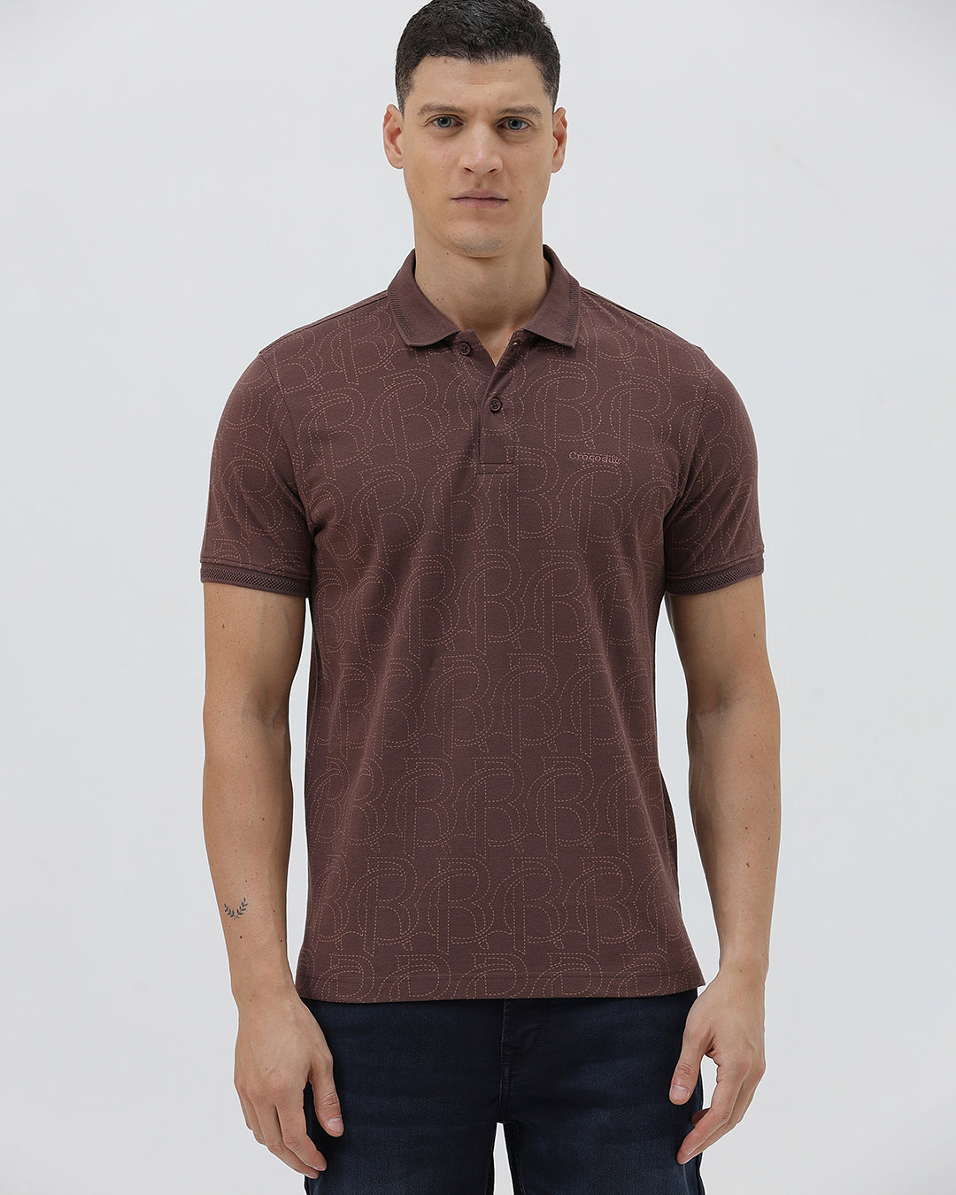 A man wearing a short-sleeve polo shirt in a dark brownish-rose color with a subtle tonal geometric pattern, paired with dark jeans, against a light gray studio background.