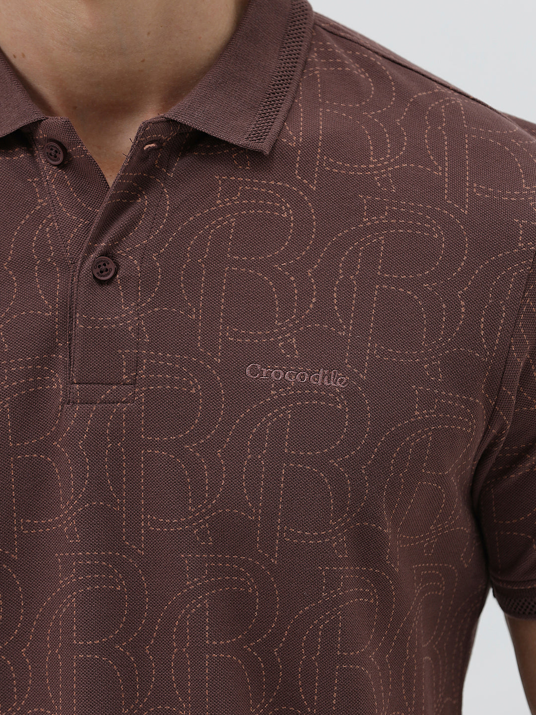 A man wearing a short-sleeve polo shirt in a dark brownish-rose color with a subtle tonal geometric pattern, paired with dark jeans, against a light gray studio background.