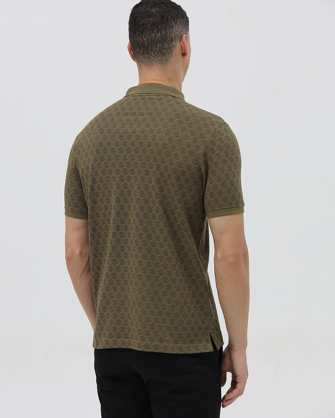 A man wearing a short-sleeve polo shirt with a subtle geometric monogram pattern in olive green and brown, paired with black trousers, against a light gray studio background.
