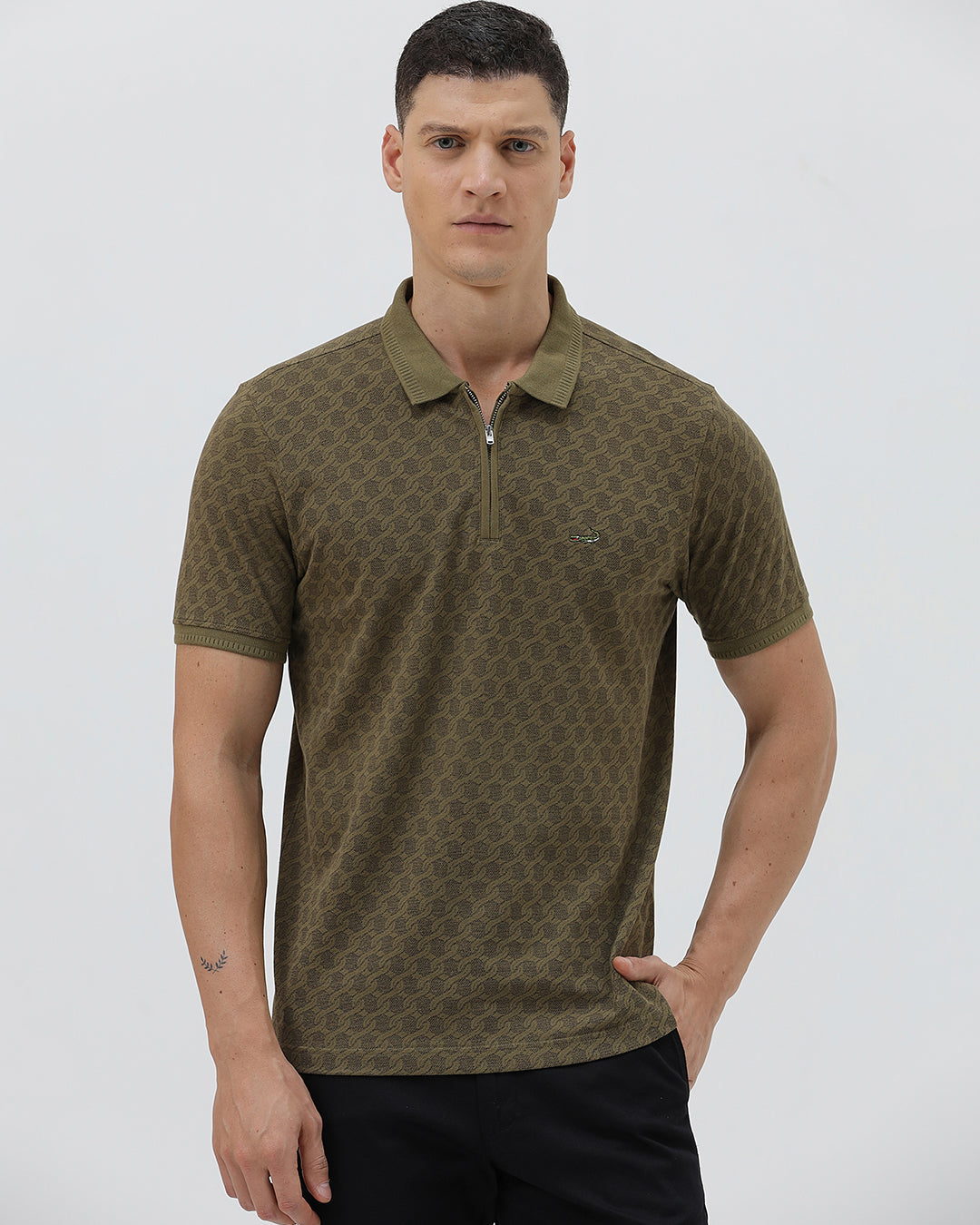 A man wearing a short-sleeve polo shirt with a subtle geometric monogram pattern in olive green and brown, paired with black trousers, against a light gray studio background.