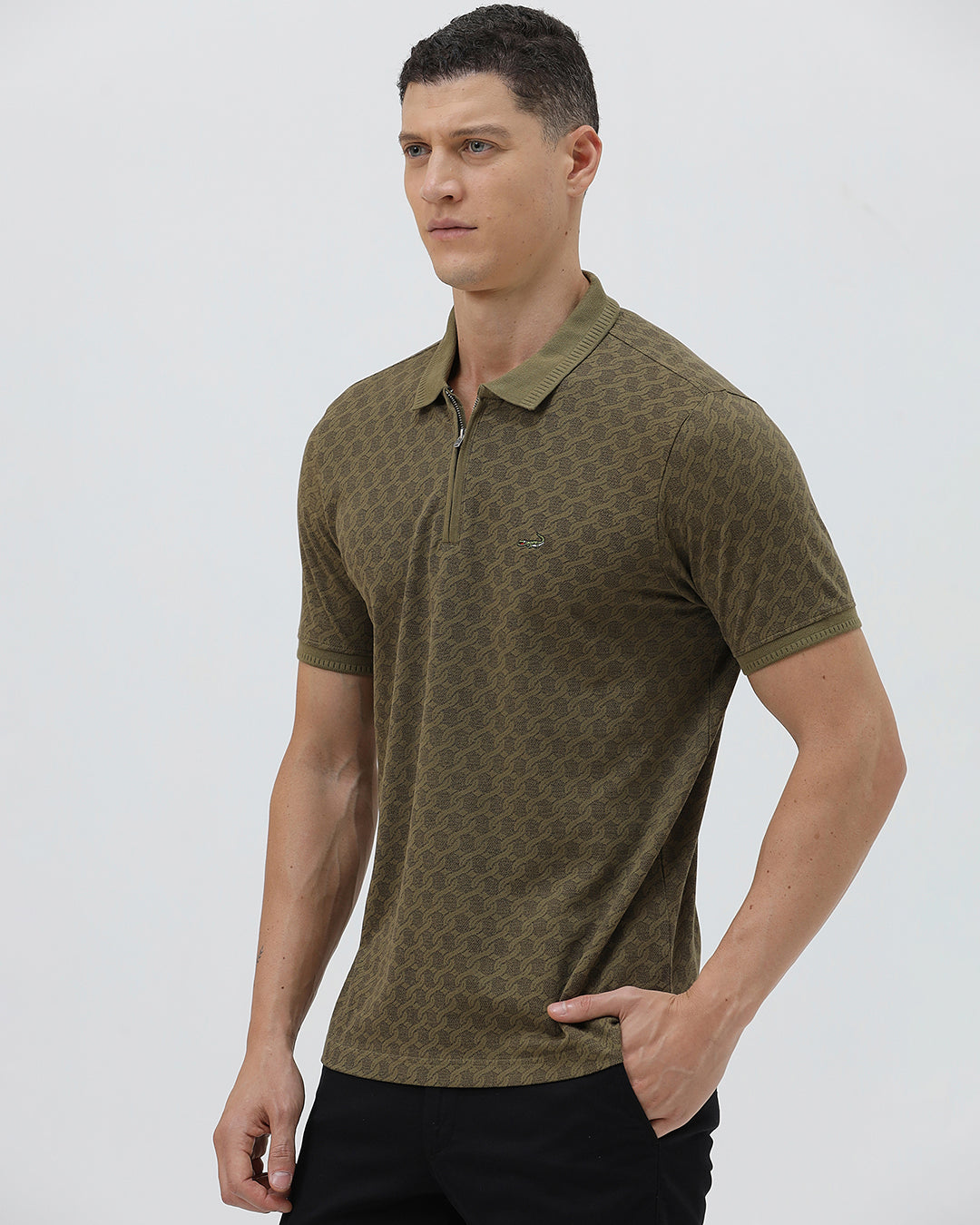 A man wearing a short-sleeve polo shirt with a subtle geometric monogram pattern in olive green and brown, paired with black trousers, against a light gray studio background.