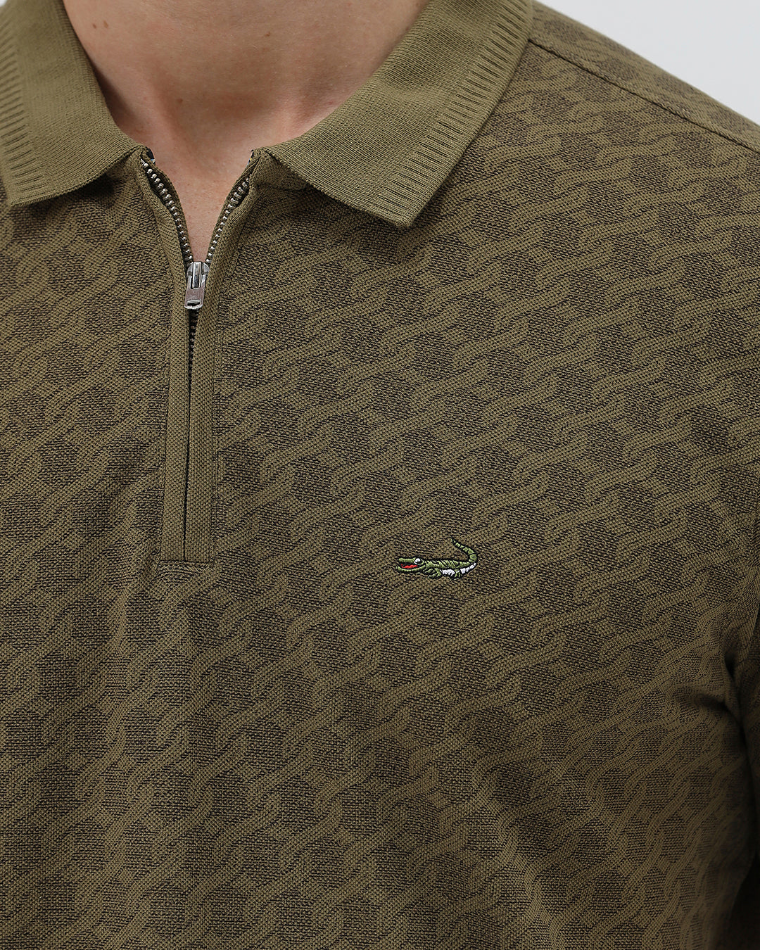 A man wearing a short-sleeve polo shirt with a subtle geometric monogram pattern in olive green and brown, paired with black trousers, against a light gray studio background.